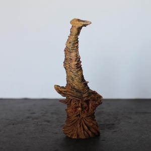 May include: A detailed, hand-carved wooden sculpture of a wizard with a long, curved staff. The sculpture is primarily brown with hints of yellow and orange, and features intricate details on the face and staff.