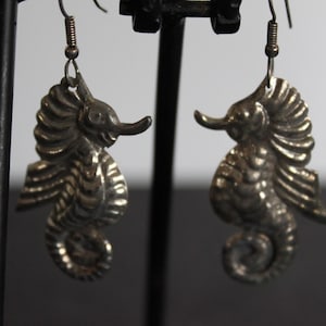 May include: A pair of silver tone earrings shaped like seahorses. The earrings are hanging from a black stand.
