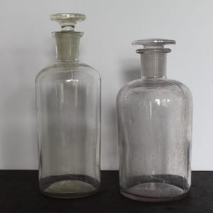 May include: Two clear glass apothecary bottles with glass stoppers. The bottles are different sizes, one tall and cylindrical, the other shorter and rounded. The stoppers have a flat, disc-shaped top. The bottles are on a dark surface.