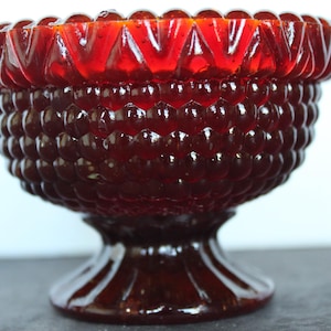 May include: A ruby red glass bowl with a textured, bubble-like pattern on the body and a decorative, geometric rim. The bowl sits on a pedestal base, showcasing a vintage aesthetic. The glass has a glossy finish.