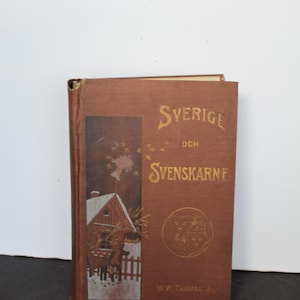 May include: A brown hardcover book titled Sverige och Svenskarne by W.W. Thomas Jr. The front cover features a gold-colored illustration of a snowy house with a chimney emitting smoke, a small fence, and a circular emblem with a crest design.