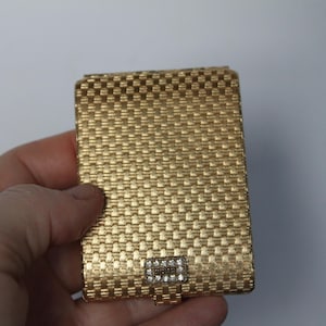 May include: A rectangular gold-toned metal cigarette case featuring a textured basketweave pattern across the surface. The case includes a small, rectangular clasp at the bottom center adorned with a row of clear, sparkling rhinestones.