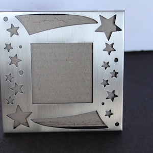 May include: A square, silver-colored metal picture frame. The frame features cut-out star shapes and curved designs. The center opening is a square shape with a neutral-colored backing.