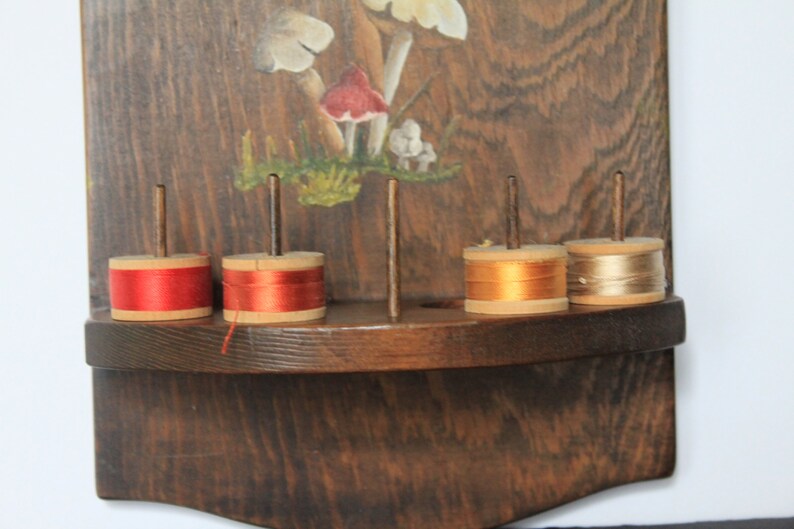 May include: Wooden wall-mounted thread rack with five spools of thread. The spools are red, orange, and beige. The rack is painted with a mushroom design.
