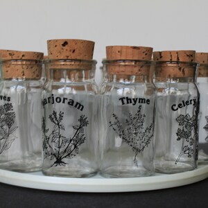 May include: A set of clear glass spice jars with cork lids, arranged on a white circular tray. Each jar is labeled with a different herb name in black, along with a corresponding botanical illustration. The herbs include cloves, marjoram, thyme, and celery.