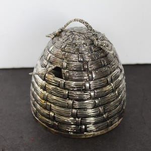 May include: Silver-colored, beehive-shaped container with a woven basket design. The container has a small opening on the side and a braided handle on top. The container is made of metal and sits on a dark surface.
