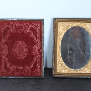 May include: Two antique daguerreotype cases. One case features a deep red velvet cover with an embossed floral pattern. The other case is open, revealing a gold-toned metal frame with an oval window containing a vintage photograph of two people.