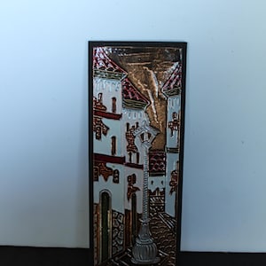 May include: A rectangular metal relief depicting a European street scene. The artwork features buildings with red roofs, a street lamp, and cobblestone streets. The color palette includes brown, white, and silver.