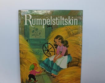 Può includere: Un libro con copertina rigida intitolato Rumpelstiltskin, A Rand McNally Giant Book. L'illustrazione di copertina raffigura una piccola figura con cappello verde e camicia rosa di fronte a una persona in camicetta rosa e gonna blu tra la paglia.