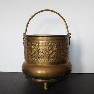May include: A vintage brass bucket with a handle. The bucket features an embossed floral design around the body and a small footed base. The handle is arched and attached to the sides. The overall color is a warm, antique gold.