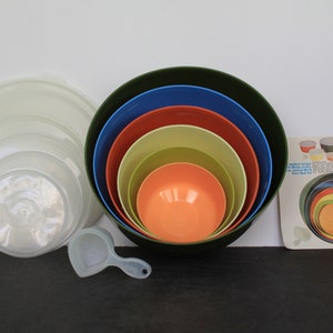 May include: A set of six nesting mixing bowls in various colors, including green, blue, orange, and yellow. The bowls are made of plastic and have a smooth, glossy finish. There are also six matching lids and a white plastic measuring cup.