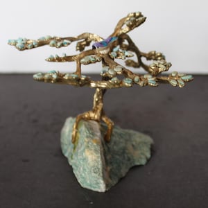 May include: A miniature brass tree sculpture with turquoise accents and a small bird perched on a branch. The tree is rooted in a green stone base, creating a decorative and artistic piece.