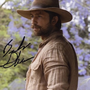 May include: A man with a mustache and beard wearing a brown hat and a light brown button-down shirt. The background is out of focus and appears to be a field of purple flowers.