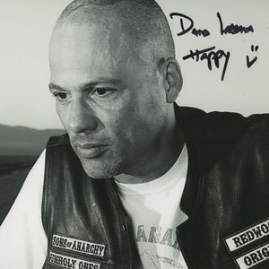 May include: Black and white photo of a man with a shaved head wearing a leather vest with patches that say "Sons of Anarchy" and "Redwood Original". The man is looking off to the side and has a serious expression on his face. The photo is signed "Dano Hazaria Happy"