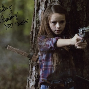 May include: A young girl in a red and blue plaid shirt holds a silver revolver in her right hand. She is standing in front of a tree with a brown leather-wrapped sword strapped to it. The girl's signature is written in black ink above the image: "Cailey Fleming XO "Judith"