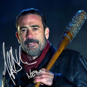 May include: A man with a beard and a red bandana around his neck is holding a baseball bat wrapped in barbed wire. The bat is brown and the barbed wire is silver. The man is wearing a black leather jacket. The man's signature is visible in the image.