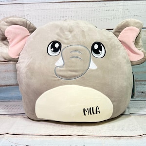 May include: A gray plush elephant pillow with pink ears and embroidered eyes. The pillow has a white patch on the bottom with the name "Mila" embroidered in black.