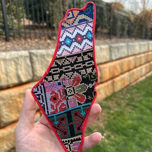 May include: A colorful, embroidered patch in the shape of a country's map. The patch features a red border and intricate patterns in various colors, including blue, pink, black, and green. The design appears to be a traditional textile pattern.