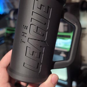 May include: A black cylindrical mug with a handle, featuring the text "THE LEAGUE" in raised lettering. The mug has a textured surface and a wide base. The mug is held in a hand.