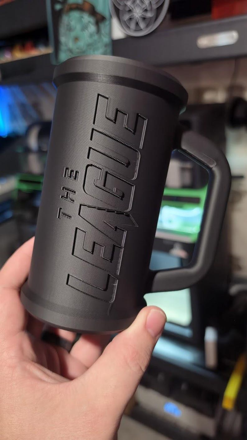 May include: A black cylindrical mug with a handle, featuring the text "THE LEAGUE" in raised lettering. The mug has a textured surface and a wide base. The mug is held in a hand.