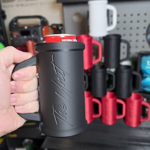 May include: Black mug with a handle, holding a red can. The mug has the text "The Wait" in a cursive font. Other mugs in red and black are stacked in the background. A Captain America shield is visible.