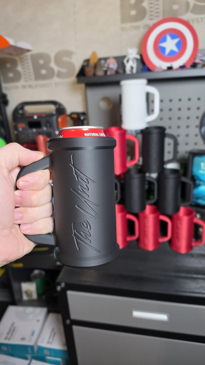 May include: Black mug with a handle, holding a red can. The mug has the text "The Wait" in a cursive font. Other mugs in red and black are stacked in the background. A Captain America shield is visible.