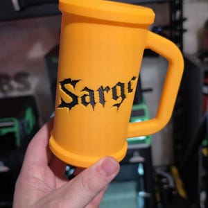 May include: An orange mug with a handle, featuring the name "Sarge" in black gothic lettering. The mug is cylindrical with a wide base and rim. A decorative emblem is visible in the background.