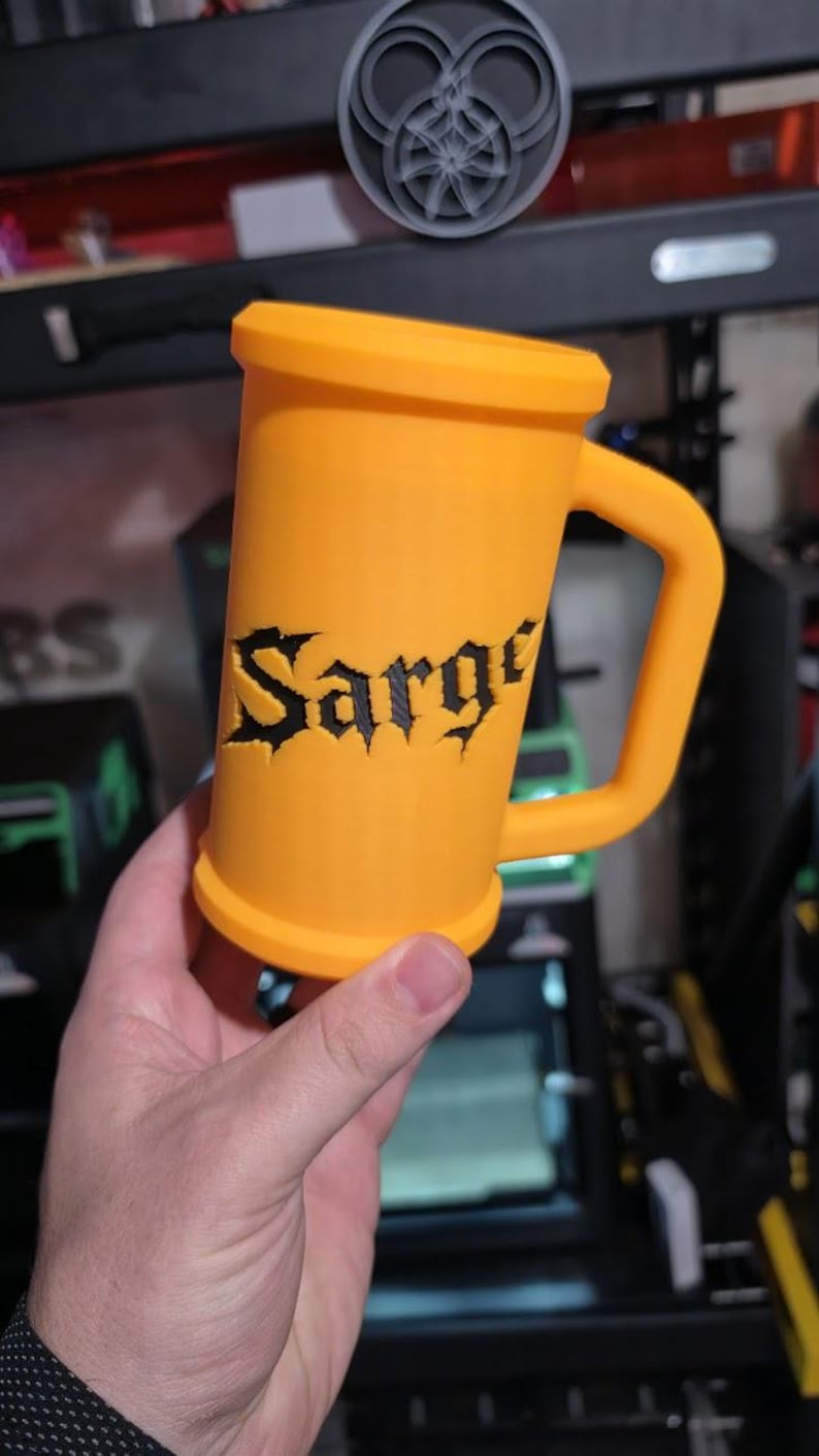 May include: An orange mug with a handle, featuring the name "Sarge" in black gothic lettering. The mug is cylindrical with a wide base and rim. A decorative emblem is visible in the background.