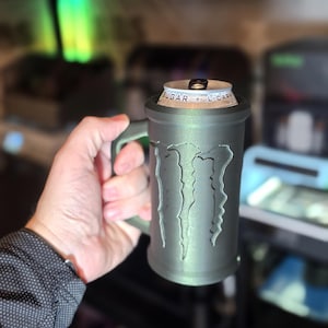 May include: A gray, 3D-printed mug with the Monster Energy logo. The mug is designed to hold a can of energy drink. The handle is integrated into the design. The can is visible at the top of the mug.
