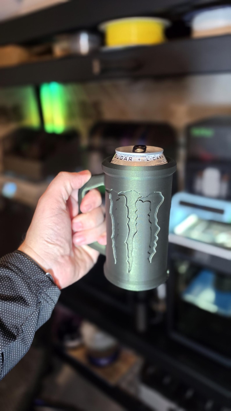 May include: A gray, 3D-printed mug with the Monster Energy logo. The mug is designed to hold a can of energy drink. The handle is integrated into the design. The can is visible at the top of the mug.