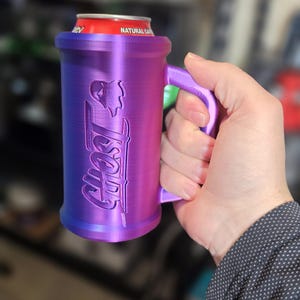 May include: A purple and blue can cooler with the word "Ghost" and a graphic of a skull. The cooler has a handle and is holding a red can. The cooler is made of plastic.