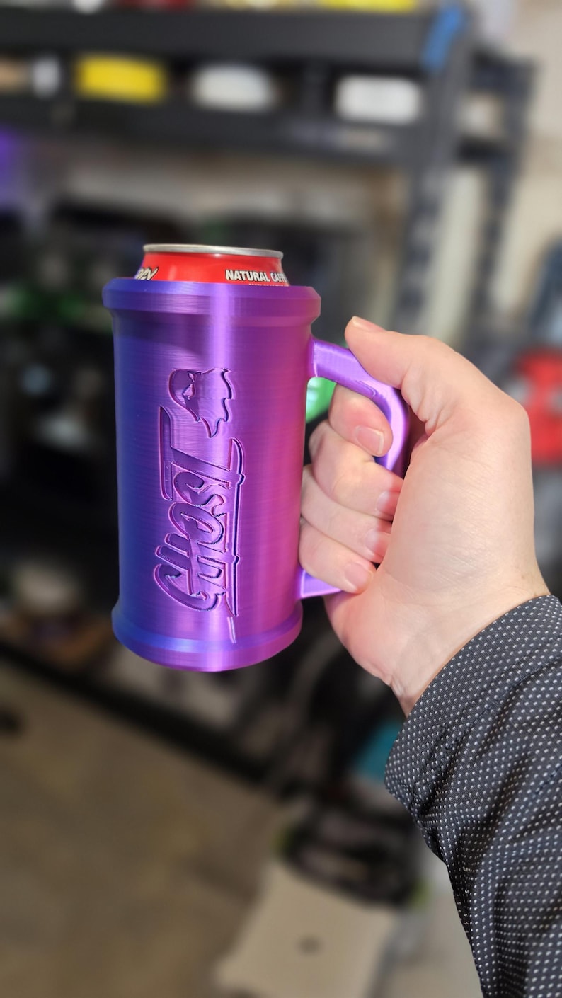 May include: A purple and blue can cooler with the word "Ghost" and a graphic of a skull. The cooler has a handle and is holding a red can. The cooler is made of plastic.