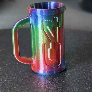 May include: A small, rainbow-colored mug with a handle. The mug has the letters "VG" embossed on the side. The colors transition from red to blue to green. The mug is made of a 3D-printed material.