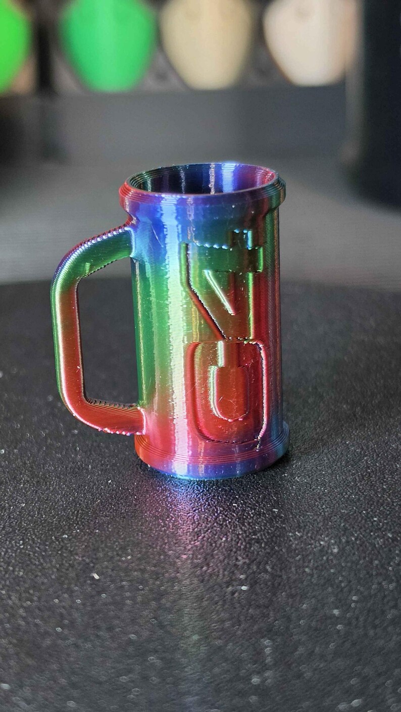 May include: A small, rainbow-colored mug with a handle. The mug has the letters "VG" embossed on the side. The colors transition from red to blue to green. The mug is made of a 3D-printed material.