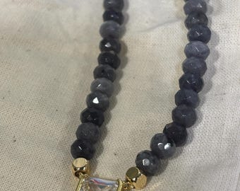 Op de afbeelding: Een ketting met facetgeslepen grijze kralen en een goudkleurige rechthoekige hanger met een heldere, vierkant geslepen steen. De ketting wordt gepresenteerd op een neutraal gekleurde stof.