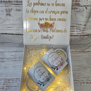 May include: A white gift box with the text "Los padrinos no se buscan" in gold lettering. Inside are two white mugs with floral designs and the words "el mejor Padrino del mundo" and "la mejor Madrina del mundo".