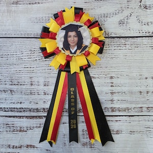 May include: A graduation rosette with a photo of a person wearing a graduation cap. The rosette features black, red, and yellow ribbons, with the text "CLASS OF 2026" on a black ribbon.