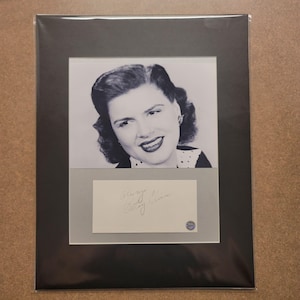 May include: A black and white portrait of a woman with dark wavy hair, set within a black mat. Below the portrait is a white rectangular card featuring the handwritten signature Always Patsy Cline and a small circular blue authentication seal.