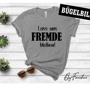 Könnte beinhalten: Graues T-Shirt mit schwarzem Text, der "Lass uns FREMD bleiben!" lautet. Der Text ist mittig auf dem Shirt platziert. Das Shirt liegt auf einem weißen Holzgrund.