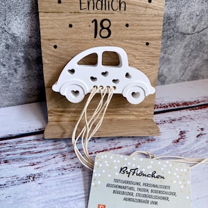 Könnte beinhalten: Holztafel mit dem deutschen Text "Endlich 18" und einem weißen Auto-Ausschnitt. Das Auto hat herzförmige Ausschnitte und ist mit Schnüren an der Tafel befestigt. Ein Anhänger mit Text und Social-Media-Symbolen hängt darunter.