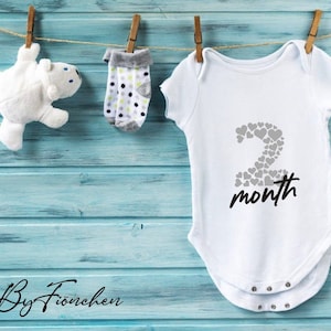 Könnte beinhalten: Weißer Baby-Strampler mit einer grauen Zahl 2 aus Herzen und dem Text "month" in Schwarz. Der Strampler hängt an einer Wäscheleine mit einem weißen Stofftier und einem Paar gepunkteten Socken.