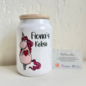 Könnte beinhalten: Weiße Keramik-Keksdose mit Holzdeckel. Die Dose zeigt ein Comic-Einhorn, das ein rotes Herz hält, und den Text "Fiona's Kekse".