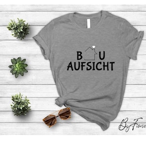 May include: Gray t-shirt with a black graphic of a house with a heart on top and the text "BAU AUFSICHT".
