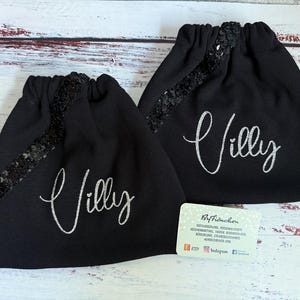 Könnte beinhalten: Zwei schwarze Kordelzugbeutel mit Paillettenakzent und dem Namen "Villy" in silberner Schrift. Die Beutel befinden sich auf einer hellen Holzoberfläche. Eine kleine Visitenkarte ist sichtbar.
