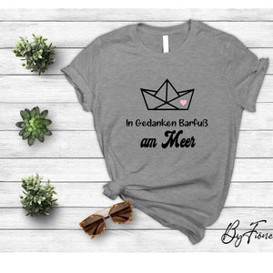May include: Gray t-shirt with a black paper boat graphic and a pink heart. The text "In Gedanken Barfuß am Meer" is printed on the shirt.