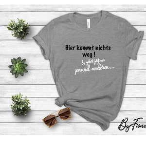 Può includere: Maglietta grigia con il testo "Hier kommt nichts weg! Es geht jetzt nur jemand anderen..." in bianco e nero. Il testo è in uno stile informale e scritto a mano. La maglietta è su uno sfondo di legno bianco.