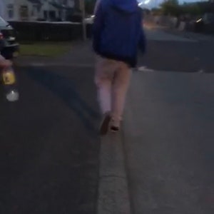 May include: A person wearing a blue jacket and light-colored pants walks down a street at night. The street is lined with houses and streetlights.