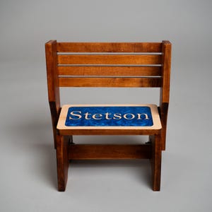 May include: A small wooden chair with a slatted backrest and a seat featuring a blue resin inlay. The name Stetson is engraved in light-colored wood lettering centered within the blue resin surface of the seat, which has a smooth finish.