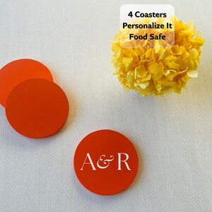 May include: Three round orange coasters, one featuring the white text A&R. A white label with black text reads 4 Coasters, Personalize It, Food Safe. The coasters are smooth and circular, presented alongside a yellow flower head.