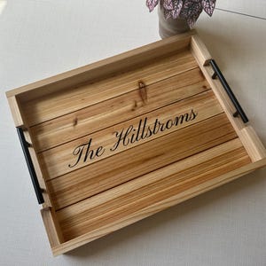 May include: A rectangular wooden serving tray featuring horizontal planks with a natural light brown grain. The tray includes two black metal handles on the sides and the text "The Hillstroms" engraved in a black cursive script in the center.